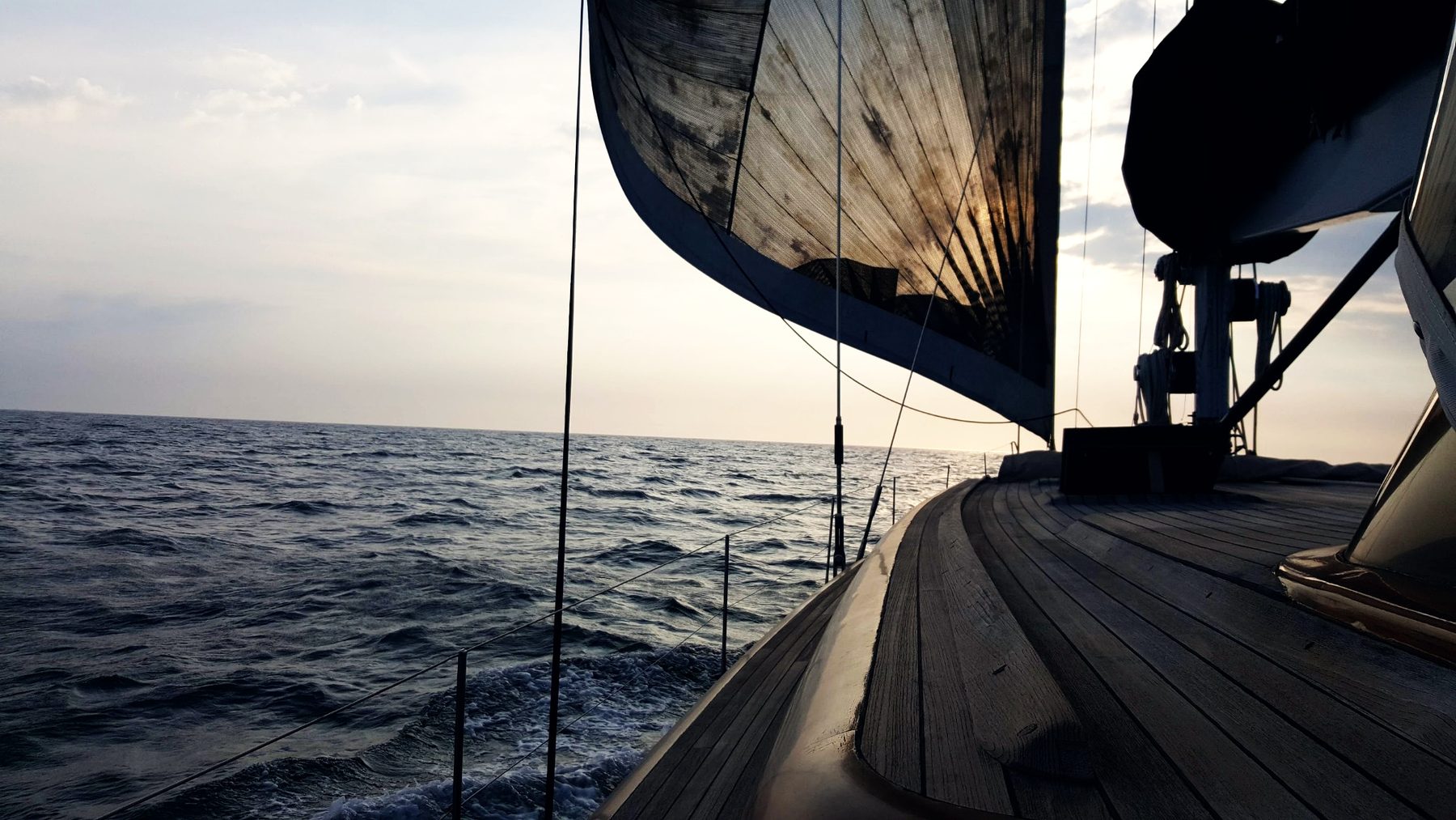 Lauren L under sail at dusk, teak side deck and headsail against a low sun
